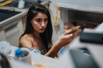 A young woman looks thoughtfully into an open refrigerator, holding a small item. The kitchen setting is modern and cozy, creating a sense of everyday life and decision-making.