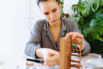 young attractive woman assembling wooden model at table in bright room with house plants