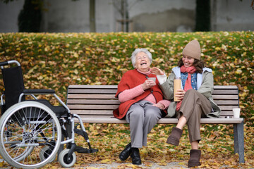 Portrait of a granddaughter and grandmother sitting on bench in autumn park, drinking coffee and laughing together.