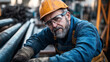 © altitudevisual - Middle-aged construction worker with gray beard wearing yellow hard hat and safety glasses working with metal pipes in an industrial setting