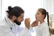 © sofiko14 - Caucasian male doctor examines throat of young girl using wooden depressor. Scene captures doctor's professionalism and care in medical setting.