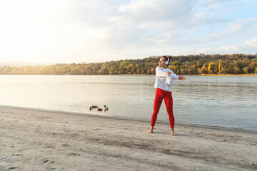 Wall Mural - Caucasian young woman in wireless headphones warming up for a workout outdoors by the riverbank in nature.