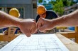 © AlAfiq - Construction Workers Celebrating a Successful Build by Fist Bumping Over Architectural Blueprint on a Sunny Day at a Job Site with Hard Hats