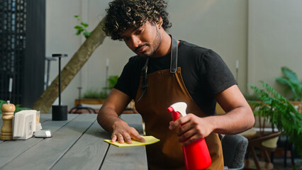 Close up smiling Latino American ethnic man male cafe worker in apron guy waiter cleaner barista wiping table in cafeteria disinfectant cleaning before opening small business street food outside city