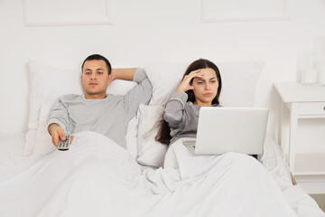 Wall Mural - Offended young woman with laptop and her husband watching TV after quarrel in bedroom