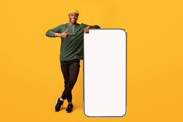 Poster - Cheerful African American Guy Leaning And Pointing At Big Smartphone With White Blank Screen, Showing Free Copy Space For Your Mobile App Or Website Design, Standing On Yellow Background, Mockup Image