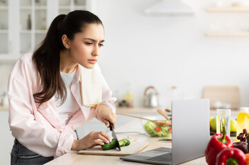 Aufkleber - Portrait of serious focused young lady cooking fresh organic salad at home in modern kitchen, using and looking at laptop on countertop, looking closer at screen, reading recipe, free copy space