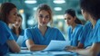 © Old Man Stocker - A group of female healthcare professionals in blue scrubs engage in a discussion, holding documents in a clinical setting, emphasizing teamwork and communication.