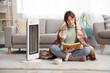 © Pixel-Shot - Young woman with warm plaid reading book near electric heater at home