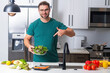 © Volodymyr - Taking care of health. Handsome man in casual t-shirt standing in the kitchen at home. Young man cooking vegetables in the kitchen. Dieting man with bowl of salad. Green salad healthy food concept.