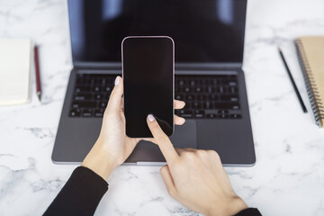 Wall Mural - Closeup view of a woman engrossed in her smartphone tasks with an open laptop nearby set against the blurred bustle of an office scene