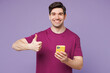 © ViDi Studio - Young smiling Caucasian man he wear violet t-shirt casual clothes hold in hand use mobile cell phone show thumb up isolated on plain pastel light purple background studio portrait. Lifestyle concept.