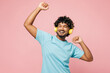 © ViDi Studio - Young cheerful fun Indian man he wear blue t-shirt casual clothes listen to music in headphones raise up hands dance isolated on plain pastel light pink background studio portrait. Lifestyle concept.