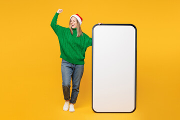 Wall Mural - Full body winner merry young woman wears green casual sweater Santa hat posing big huge blank screen mobile cell phone isolated on plain yellow background. Happy New Year, Christmas holiday concept.