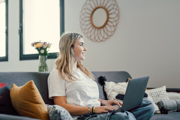 Wall Mural - Portrait of one young attractive blonde woman using laptop pc computer on couch relaxing surfing the net at home looking at the window waiting package.