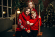 © VlaDee - Mom, son and daughter in red sweaters at home during the Christmas holidays. Beautifully decorated house in Christmas decor.