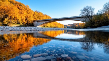  An autumn scene with golden trees reflecting in a river as a concrete bridge spans the waters, creating a mirror image of the picturesque landscape around it.