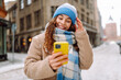 © maxbelchenko - Young woman is walking in city and viewing social nets in smartphone, chatting online and smiling. Winter weather.  Lifestyle, travel, tourism, technology