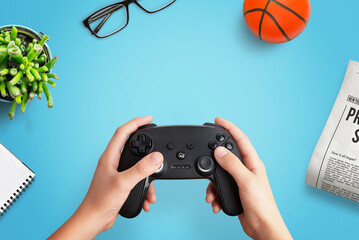 Poster - Top view flat lay composition of a player holding a gamepad on a desk with a ball, newspaper, glasses, plant, and notepad, perfect for creative gaming themes