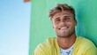 © Lens Legacy - A cheerful young man with wavy hair stands smiling confidently against a vibrant green wall, dressed in a casual yellow shirt, embodying a joyful and relaxed mood.