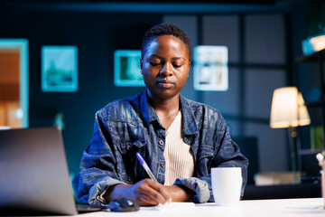Poster - Young black woman studying while using her laptop and taking notes on a notepad. African American female entrepreneur browsing the internet and writing down new business ideas.