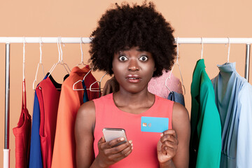 Poster - Mobile Shopping App. Shocked African American Woman Holds Mobile Phone And Credit Card For Online Shopping Looking At Camera Posing Near Clothing Rail On Yellow Background. Studio Shot