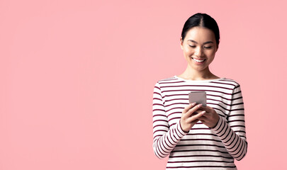 Wall Mural - Mobile Communication. Young Happy Asian Woman Using Modern Smartphone For Messaging With Friends, Smiling Korean Female Typing On Cellphone While Standing Over Pink Studio Background, Free Space