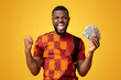 © Prostock-studio - Happy african american guy in traditional t-shirt holding bunch of money and shouting, yellow background