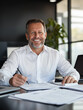 © Mavita - charismatic businessman sitting at sleek office desk, smiling confidently while reviewing documents. modern workspace features laptop and greenery, creating professional atmosphere