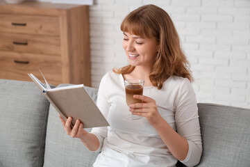 Poster - Happy young woman with glass of tasty iced latte and book sitting on sofa at home