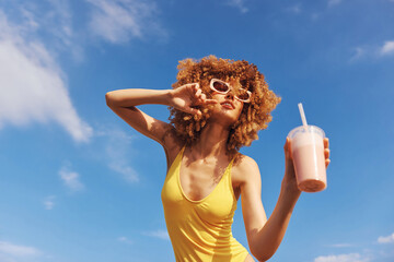 Young woman enjoying a smoothie against a blue sky background She poses confidently, wearing stylish sunglasses and a yellow swimsuit