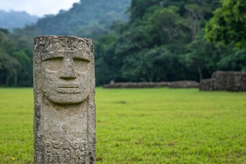 Wall Mural - Ancient stone idol statue in archaeological park with carved face. Pre-Columbian artifact on green grass field against tropical forest background. Historical heritage site with copy space