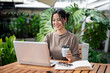 © bongkarn - A charming Asian woman working remotely from a serene green cafe, enjoying a coffee while working.