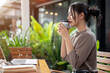 © bongkarn - A beautiful, calm Asian woman enjoys a hot drink while working remotely from a coffee shop.