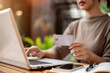 © bongkarn - A woman holding a credit card and using her laptop while sitting at an outdoor table of a cafe.