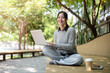 © bongkarn - Happy Asian woman works remotely outdoors, sitting on a park bench with her laptop, wears earphones.