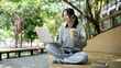© bongkarn - A woman enjoys a hot drink while working remotely outdoors, sits on a park bench with her laptop.