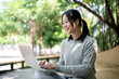 © bongkarn - A charming Asian woman works remotely outdoors, sitting on a park bench with her laptop.