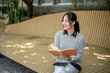 © bongkarn - A woman enjoys listening to music through her earphones and reading while sitting on a park bench.