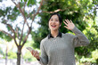© bongkarn - A cheerful Asian woman waves to greet her friends while walking along the sidewalk in a park.