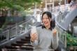 © bongkarn - A cheerful woman sits outdoors, holding up a victory fist with a big smile as she looks at her phone