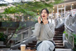 © bongkarn - A cheerful woman sits outdoors, listens to a surprising story during a fun conversation on the phone