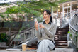 © bongkarn - A beautiful, happy Asian woman sits outdoors on a bright day, using her smartphone with a smile.