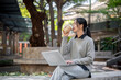 © bongkarn - A charming Asian woman enjoys a hot drink while working on her laptop outdoors.