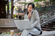 © bongkarn - A charming Asian woman sitting outdoors, talking on the phone while working on her laptop.