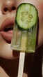 © RaptorWoman - Close-up of a woman's mouth next to a cucumber popsicle, highlighting refreshing and summery vibes.