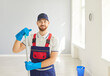 © Studio Romantic - Happy smiling male young janitor in uniform from professional cleaning service looking cheerful at camera indoors. Man cleaner holding mop washing floor in empty room or at office.