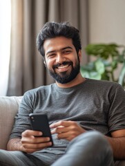 Happy young indian man using smart phone sitting on couch at home. Smiling bearded ethnic guy holding mobile phone watching social media video on smartphone, ordering food delivery online or messagin