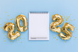 © olyphotostories - Flat lay of a blank spiral notebook with a pen, surrounded by shiny golden 2025 foil balloons on a pastel blue background. Perfect for New Year resolutions, planning, or celebration themes