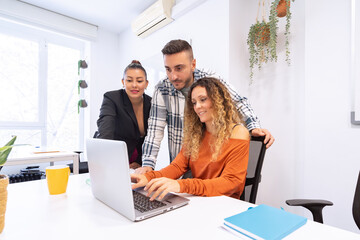 a multi-ethnic team of three collaborates around a laptop in a bright office adorned with green plan